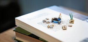 En modell av en människa i miniatyr som sitter vid ett skrivbord uppställt ovanpå några böcker som ligger i en hög